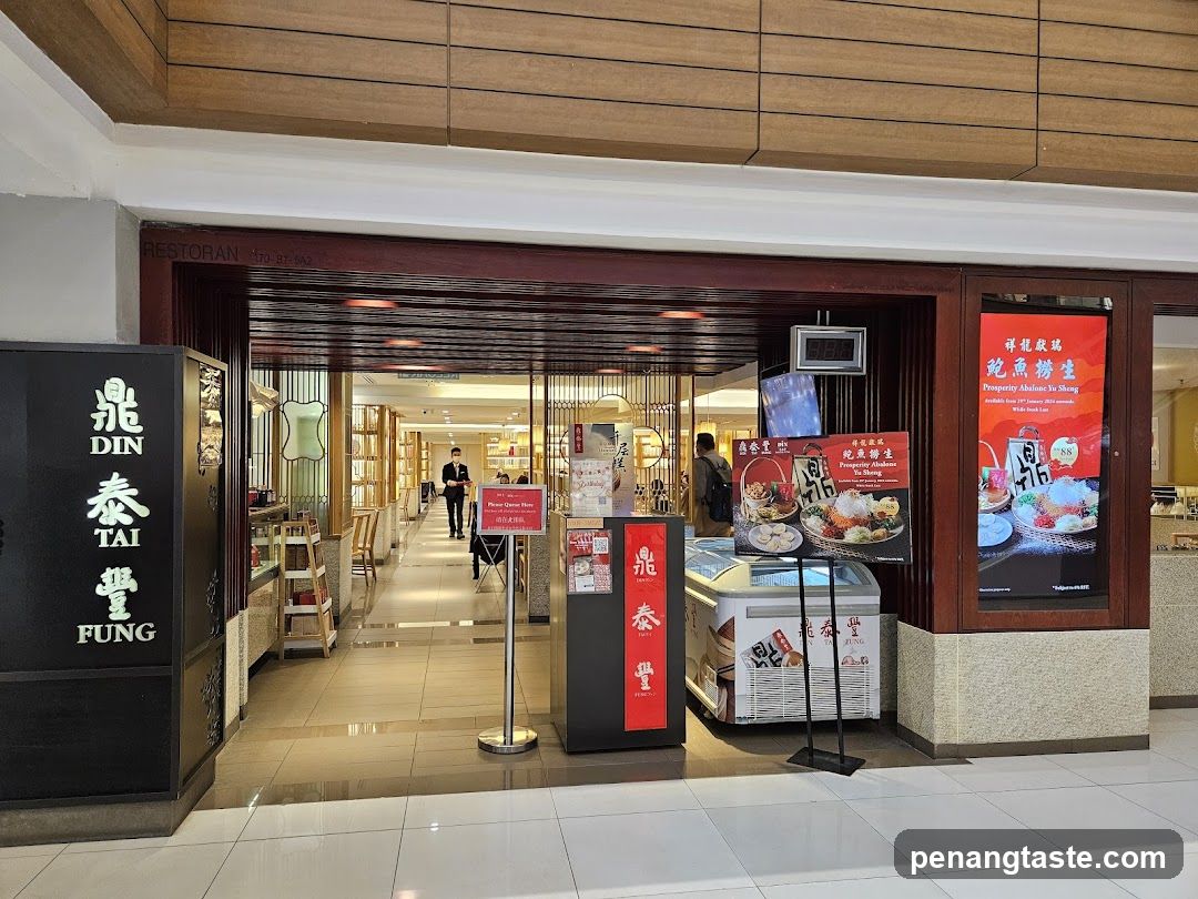 The exterior of a Din Tai Fung restaurant. A large black sign with the restaurant's name in Chinese characters and English is visible on the left. The entrance is wide, with a dark red wooden frame. Inside, the restaurant appears well-lit with tables and chairs. A staff member in uniform is visible walking inside. Promotional posters for "Prosperity Abalone Yu Sheng" are displayed near the entrance.