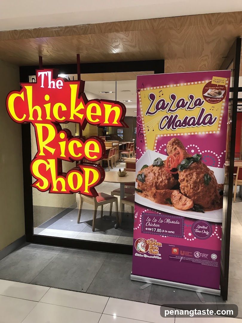 A brightly lit sign for "The Chicken Rice Shop" hangs above the entrance to the restaurant. To the right of the entrance, a promotional banner displays an image of "La La La Masala Chicken" and its price.