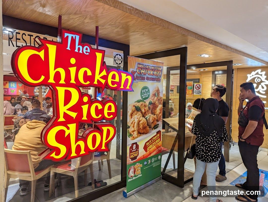 A brightly lit storefront with a large red and yellow sign that reads "The Chicken Rice Shop". Inside, customers are seated at tables. A promotional poster for grilled chicken is displayed near the entrance.