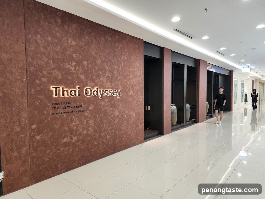 The exterior of a Thai massage parlor called Thai Odyssey. The sign is illuminated and located on a textured brown wall. Below the name, it reads "PURE MASSAGE, TIMELESS TRADITION" and some text in Thai. The entrance appears to be a series of dark doorways with decorative vases inside. A person is walking past the entrance in a mall corridor.