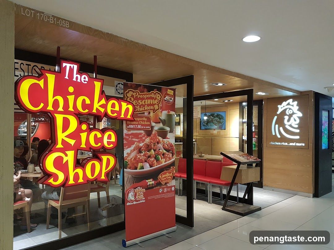 The exterior of a restaurant called The Chicken Rice Shop. A large red and yellow sign with the restaurant's name is prominently displayed. To the right, a white chicken logo with the text "chicken rice...and more" is visible. A promotional banner for "Prosperity Sesame Chicken" is also present.