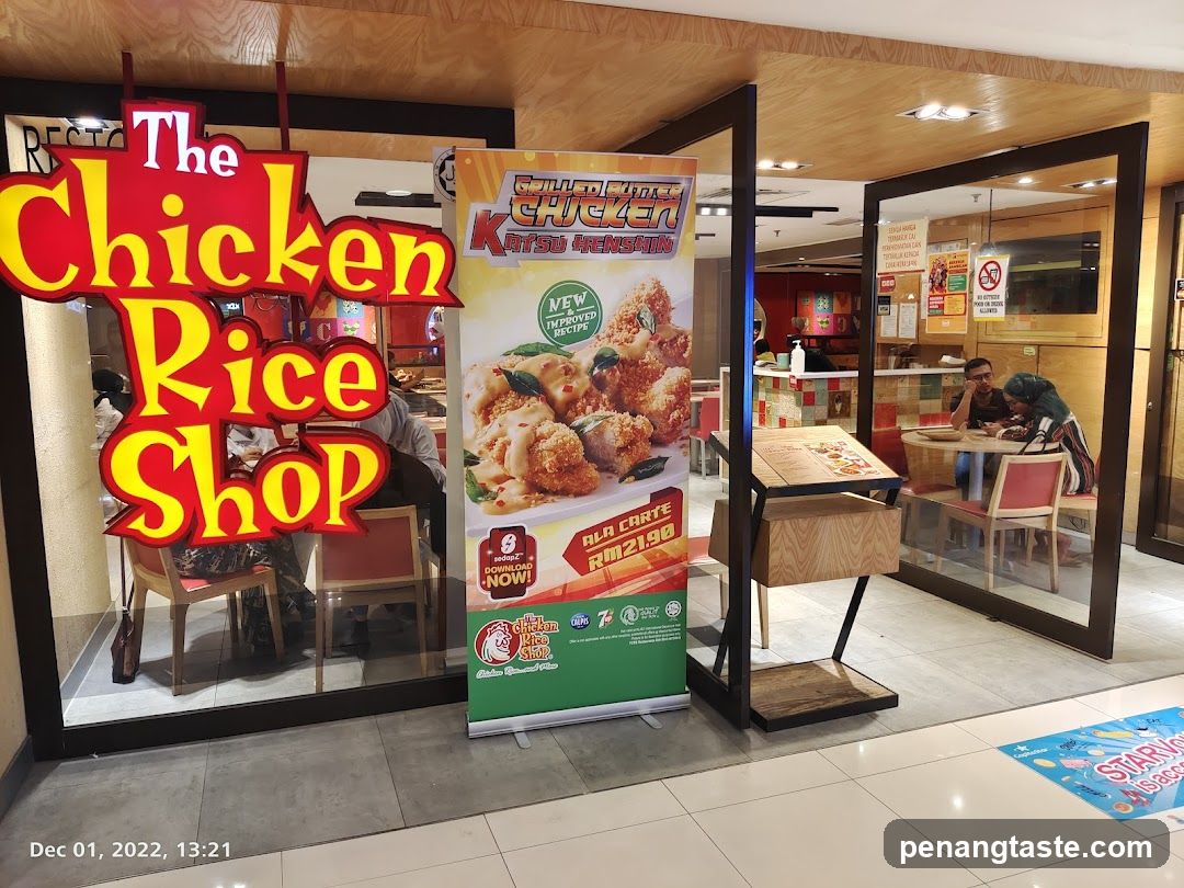 The exterior of a restaurant called "The Chicken Rice Shop". A large red and yellow sign with the shop's name is visible. A promotional banner for "Grillout Chicken Katsu Henshin" is displayed outside the entrance. Inside, customers are seated at tables.