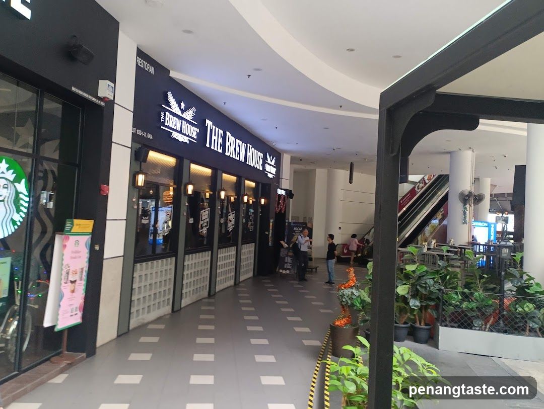 The exterior of a restaurant called The Brew House, with its name prominently displayed on a lit sign above the entrance. The facade features large windows and a black and white color scheme. To the left, a Starbucks sign is visible. The walkway in front of the restaurant is paved with a checkered pattern. In the background, escalators and seating areas can be seen within the mall.