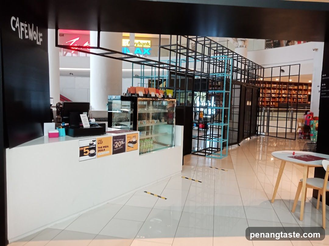 The exterior of a cafe with a black facade and a white counter. The cafe's name, "CAFE WOLF", is visible on the black wall. There are promotional signs on the counter advertising meal deals and discounts. Behind the counter, a display case shows pastries. The interior features a modern black metal shelving unit and a round table with chairs. In the background, a bookshelf is visible.