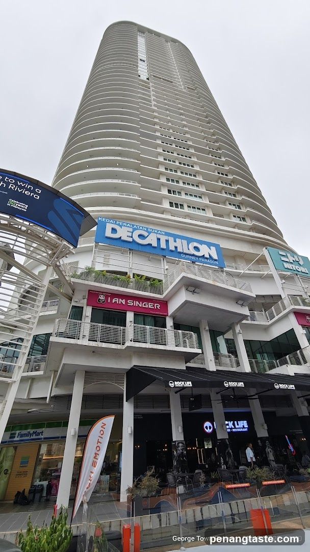 A low-angle shot of a tall, modern building with multiple levels of shops. The prominent signage for 'DECATHLON' is visible on the upper level, along with 'NITORI' and 'I AM SINGER' on lower levels. Below these, 'FamilyMart' and 'ROCK LIFE' (a cafe/restaurant) are also visible. The building has a distinctive curved facade with many balconies.