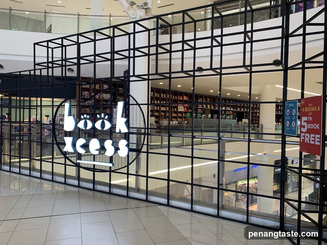 The exterior of a bookstore with a large circular sign displaying the store name 'Book Xcess' in white lettering. The store is visible through a black metal grid, showing shelves filled with books and a counter area. A promotional sign is visible on the right, advertising 'Buy 4 Books & Get 5th Book FREE'.