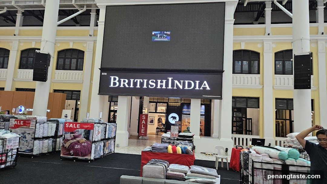 The storefront of a British India store is visible, with a large illuminated sign displaying the store name. Below the sign, the entrance to the store is open, revealing displays of clothing and other merchandise. In front of the store, there are racks of bedding and pillows, some marked with "SALE" signs. A large digital screen is mounted above the store sign.