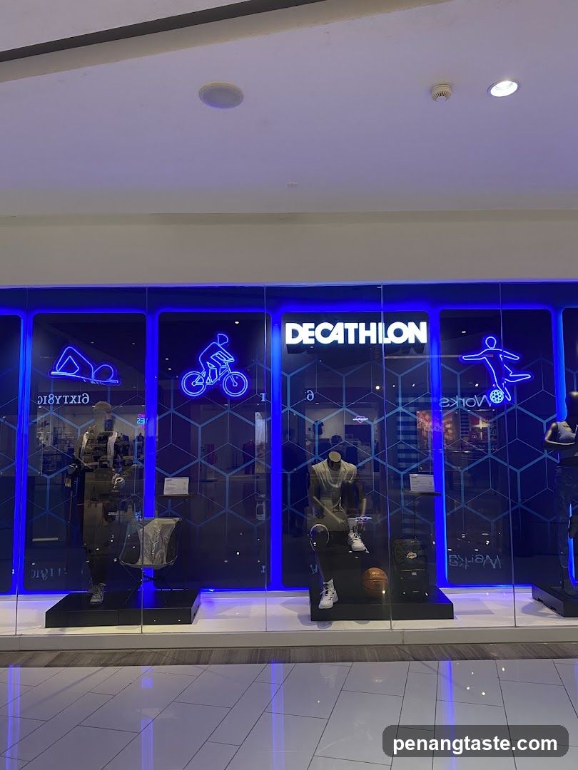 The storefront of a Decathlon store is visible, featuring large glass windows and bright blue neon lighting. The store's name, "DECATHLON," is prominently displayed in white lettering above the windows. Inside the windows, mannequins are displayed wearing athletic apparel and shoes, with sports equipment like a basketball and backpacks also showcased. Neon outlines of figures engaged in sports like cycling and yoga are visible on the glass.