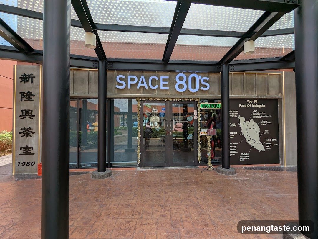 Exterior view of a modern storefront with black metal framework and perforated glass roof. The shop name 'SPACE 80's' is displayed in large white letters above the entrance. To the left, a vertical stone plaque reads '新隆興茶室 1980'. A sign on the right lists 'Top 10 Food Of Malaysia' with a map of Malaysia. Glass doors are decorated with gold tinsel. A green sign says 'NO PORK NO LARO'.