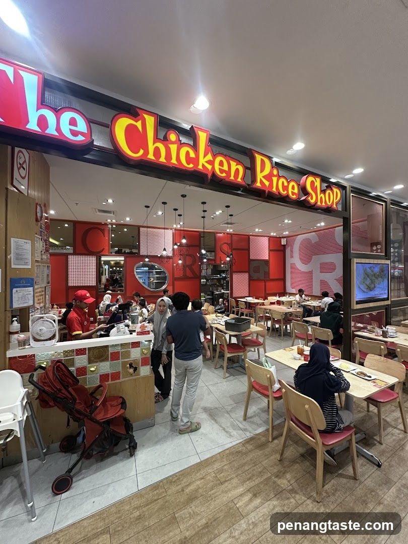 Storefront with bright red and yellow neon sign reading 'The Chicken Rice Shop'. Interior visible through glass doors; customers seated at wooden tables with red cushions. Staff in red uniforms are present near counter. A stroller is parked outside the entrance. The floor transitions from tiled to wood laminate. Lighting fixtures hang above dining area.