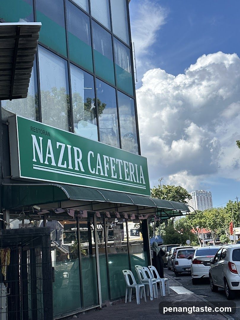 A photo of the physical storefront facade showing a green sign with white text reading 'RESTORAN NAZIR CAFETERIA' mounted on a glass building. Below, there is an awning and several white plastic chairs placed outside near parked cars under a partly cloudy sky.