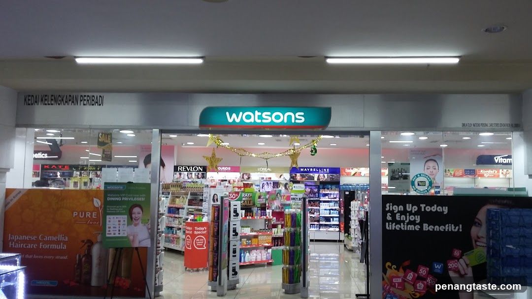 A brightly lit storefront of a Watson's pharmacy and beauty store, featuring the brand name prominently displayed above glass doors. Inside, shelves are stocked with various products including Revlon, Maybelline, and other cosmetic brands. Promotional banners advertise Japanese Camellia Haircare Formula and lifetime benefits for signing up.