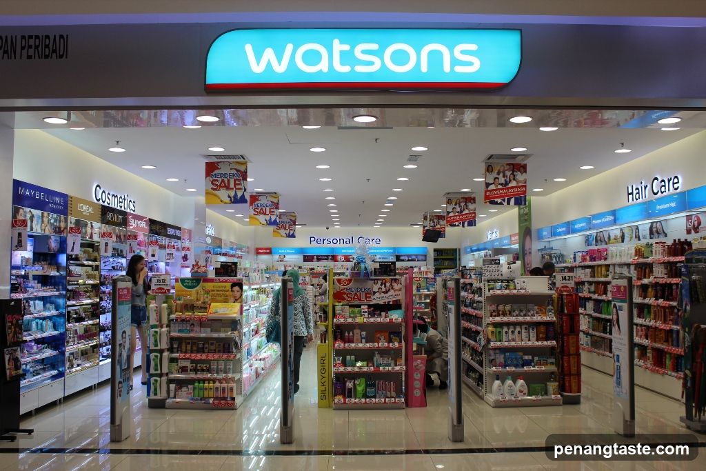 The image shows the storefront of a Watson's pharmacy and beauty store, with its illuminated blue sign prominently displayed above the entrance. Inside, various product sections are visible, including Cosmetics, Personal Care, and Hair Care. Shelves are stocked with an array of products, and promotional banners advertise sales.