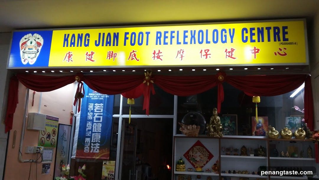 A storefront with a yellow signboard displaying 'KANG JIAN FOOT REFLEXOLOGY CENTRE' in English and Chinese characters. Red drapes hang above the entrance, adorned with decorative elements. Inside, shelves display various items including statues and figurines.