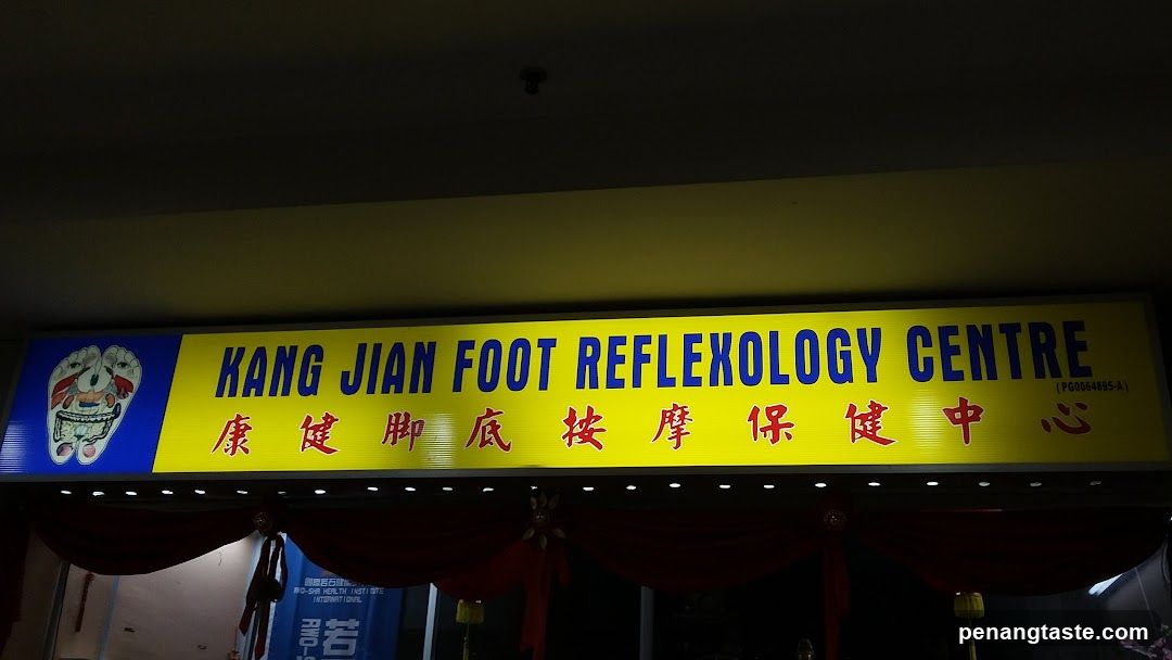 A brightly lit storefront sign with yellow background and blue text reading 'KANG JIAN REFLEXOLOGY CENTRE' in English, accompanied by Chinese characters below. A logo of a foot is visible on the left side. Red drapes hang beneath the sign.