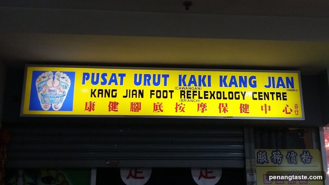 A brightly lit yellow storefront sign with blue and red text, featuring both English and Chinese characters. The sign reads 'PUSAT URUT KAKI KANG JIAN' and 'KANG JIAN FOOT REFLEXOLOGY CENTRE', along with a diagram of foot reflexology points.