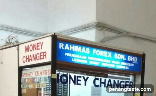 A shop front with signage reading 'Money Changer' and 'Rahimas Forex Sdn Bhd'. The sign also mentions 'Permanahan Perniagaan' and other text in Malay. Below the main sign, there is a display of currency notes.