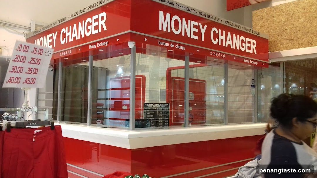 A red and white money exchange booth with visible signage reading 'MONEY CHANGER' in English, French ('bureau de change'), and Chinese characters. A price list is displayed on the left side showing rates like 10.00 to 45.00.