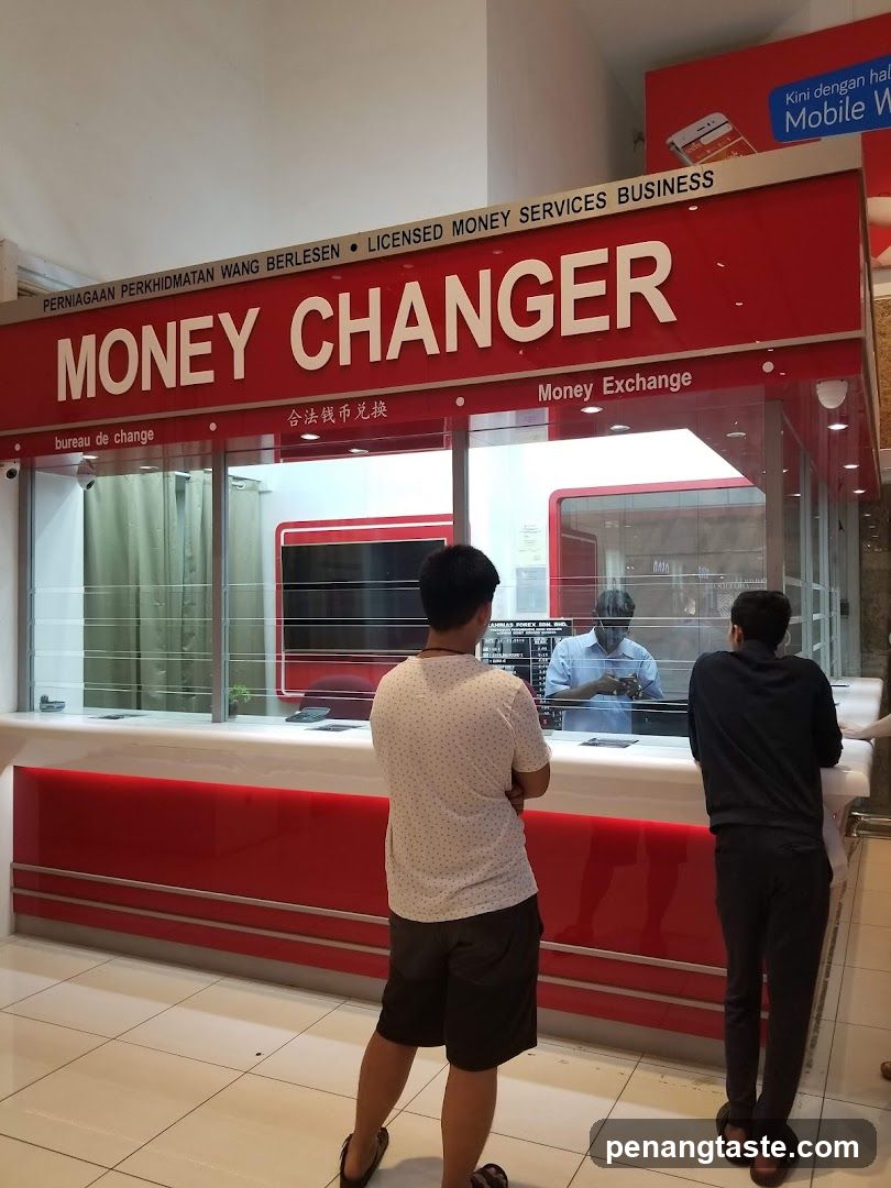 A red and white money changer storefront with visible signage reading 'MONEY CHANGER' in English, Chinese ('合法钱币兑换'), and French ('bureau de change'). The shop is licensed as a 'LICENSED MONEY SERVICES BUSINESS'. Two customers stand at the counter facing an employee behind glass.