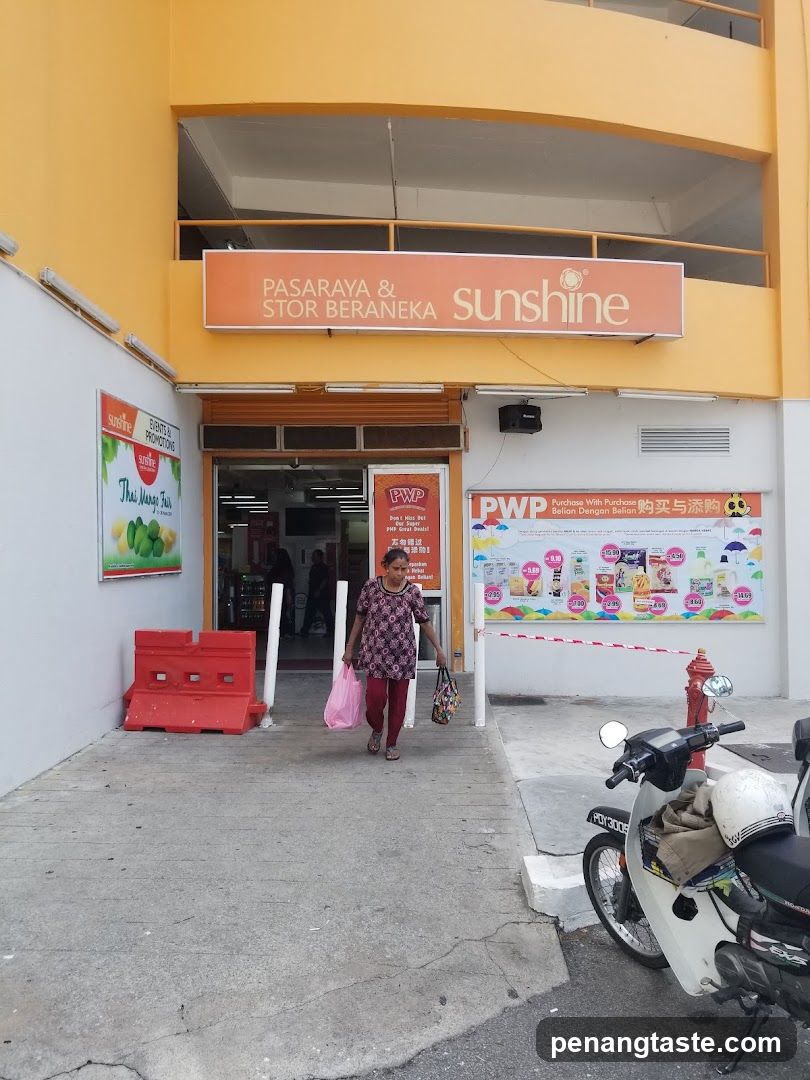 Exterior view of a supermarket entrance with orange and white facade. A woman exits carrying shopping bags. Red barriers on left, motorcycle parked right. Signage reads 'PASARAYA & STORE BERANEKA sunshine'. PWP promotional banner visible near door.