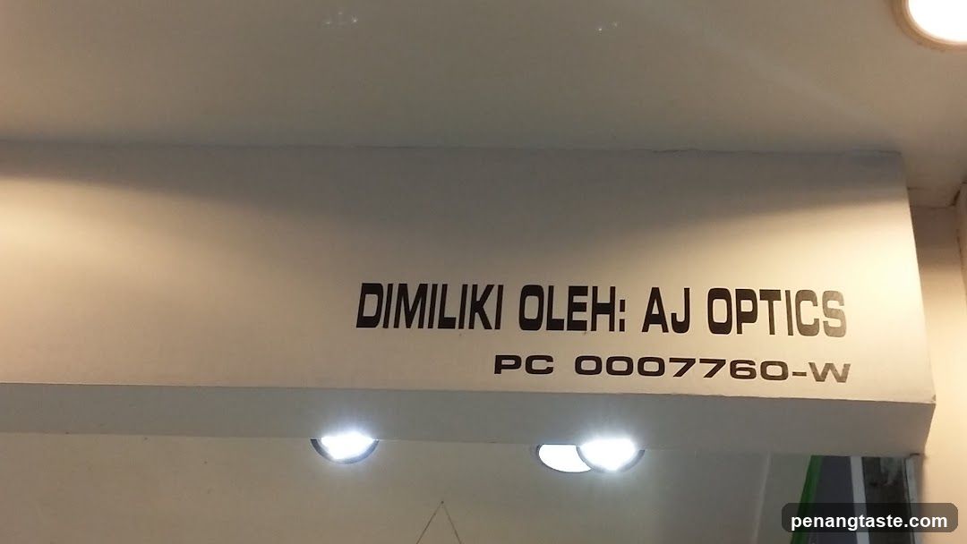 The image shows a shop front with signage that reads 'DIMILIKI OLEH: AJ OPTICS' and includes the registration number 'PC 0007760-W'. The sign is mounted on a white wall above the entrance, illuminated by ceiling lights.