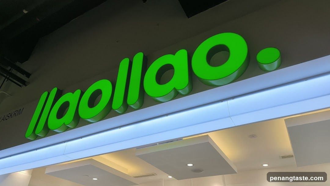A close-up view of a brightly lit storefront featuring large, three-dimensional green lettering spelling 'llaollao.' mounted on the building facade above an illuminated white beam. The sign is backlit and casts a glow onto the ceiling below. Partially visible to the left is smaller text reading 'AISKRIM'. The interior ceiling shows recessed lighting panels.