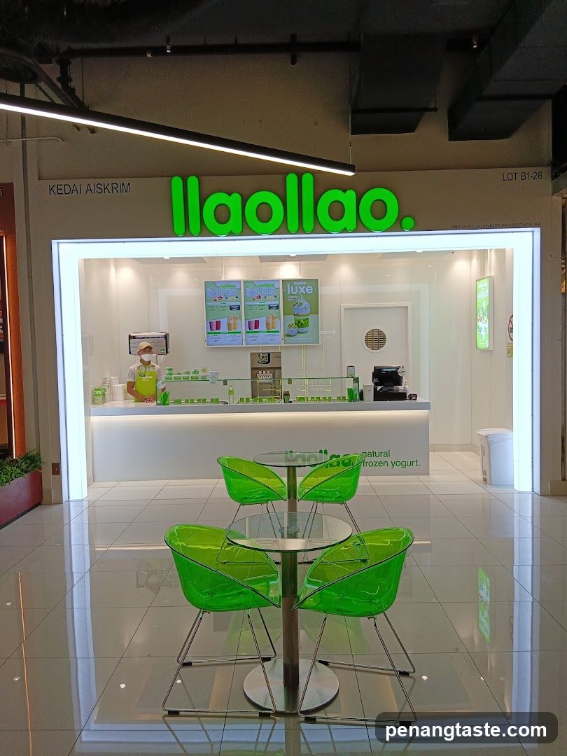 A brightly lit storefront of 'llaollao' with green neon signage above the entrance. Inside, a staff member in yellow apron stands behind the counter displaying various frozen yogurt products and drinks. A glass table with four translucent green chairs sits outside near the entrance.