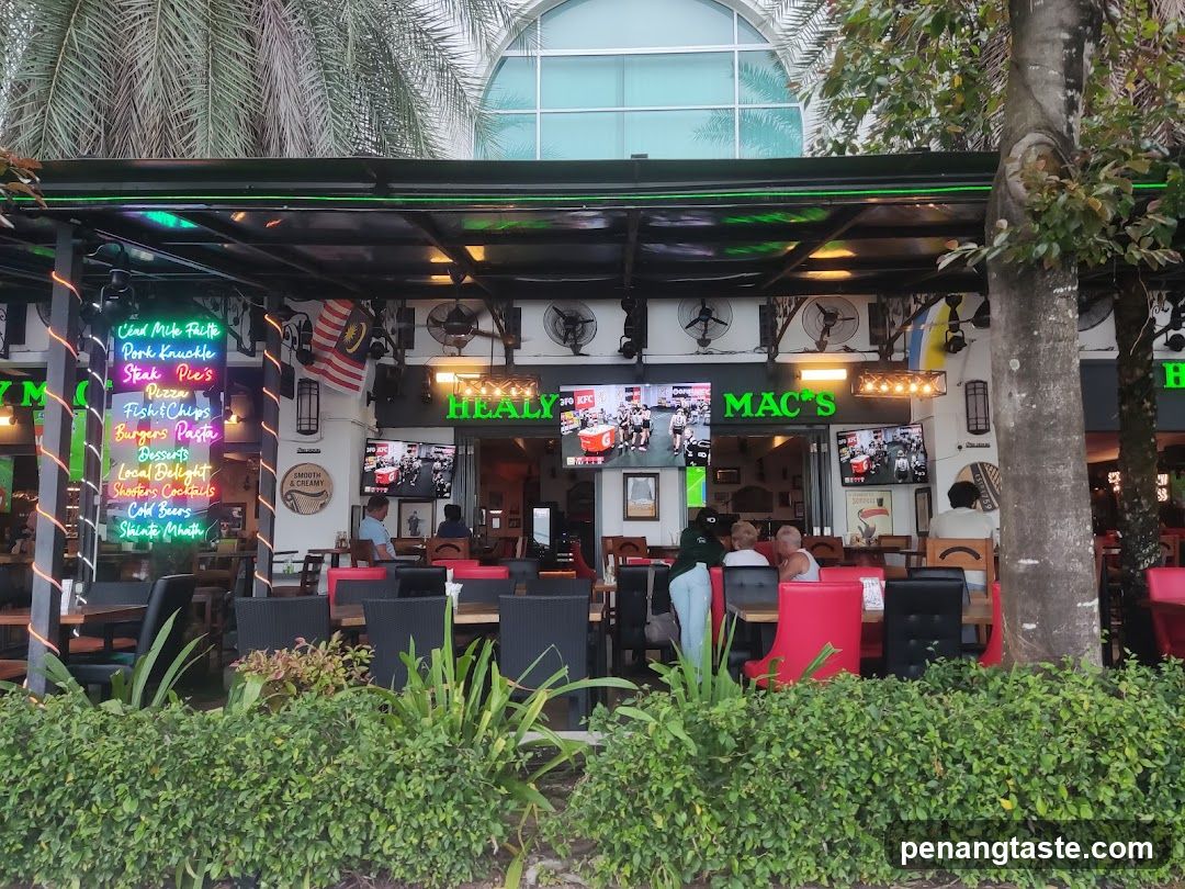 A restaurant exterior featuring a black awning, neon signs reading 'HEALY' and 'MAC'S', multiple TV screens showing food content, and a vertical menu board listing items like 'Vindaloo Pork Knuckle', 'Steak Pie's', 'Fish & Chips', etc. The scene includes outdoor seating with red chairs, greenery in the foreground, palm trees above, and visible Malaysian flags.