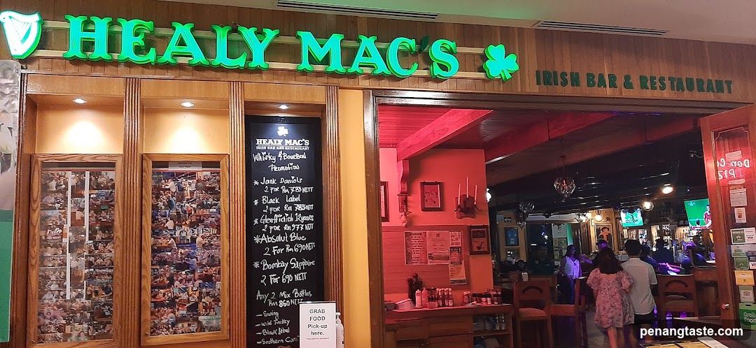 The image shows the storefront of Healy Mac's, an Irish bar and restaurant. The sign is illuminated in green with shamrocks on either side. There are two framed photo collages to the left of the entrance. A chalkboard menu board lists various drinks and prices. Inside, there are tables, chairs, and people dining.