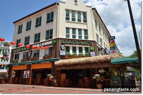 A corner building with a pub and restaurant facade, featuring green signage reading 'HEALY MAC'S PUB & RESTAURANT' and 'HEALY MAC'S'. Red lanterns hang above the entrance. A thatched roof awning covers an outdoor seating area with wooden tables and chairs. Potted plants are placed around the entrance. The building has multiple stories with large windows.