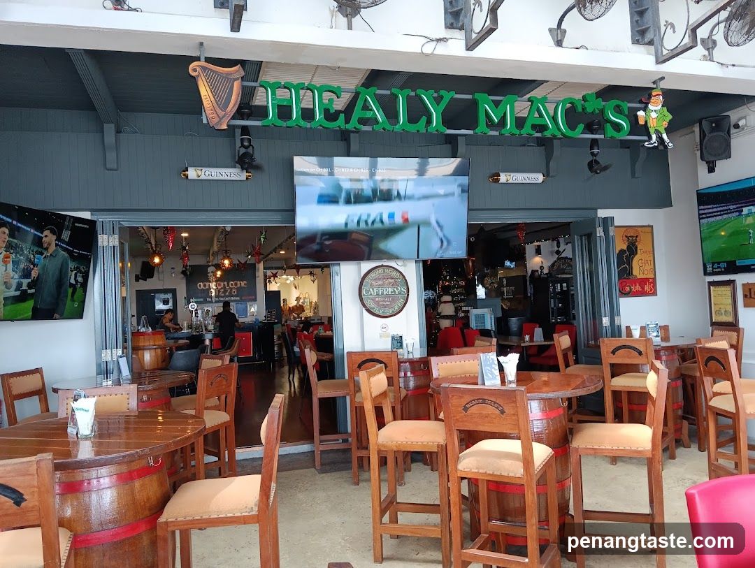 The image shows the exterior of Healy Mac's pub, featuring a prominent green sign with 'HEALY MAC'S' and a harp logo above the entrance. The facade includes multiple screens displaying sports content, wooden tables and chairs arranged outside, and visible interior elements like bar counters and patrons.