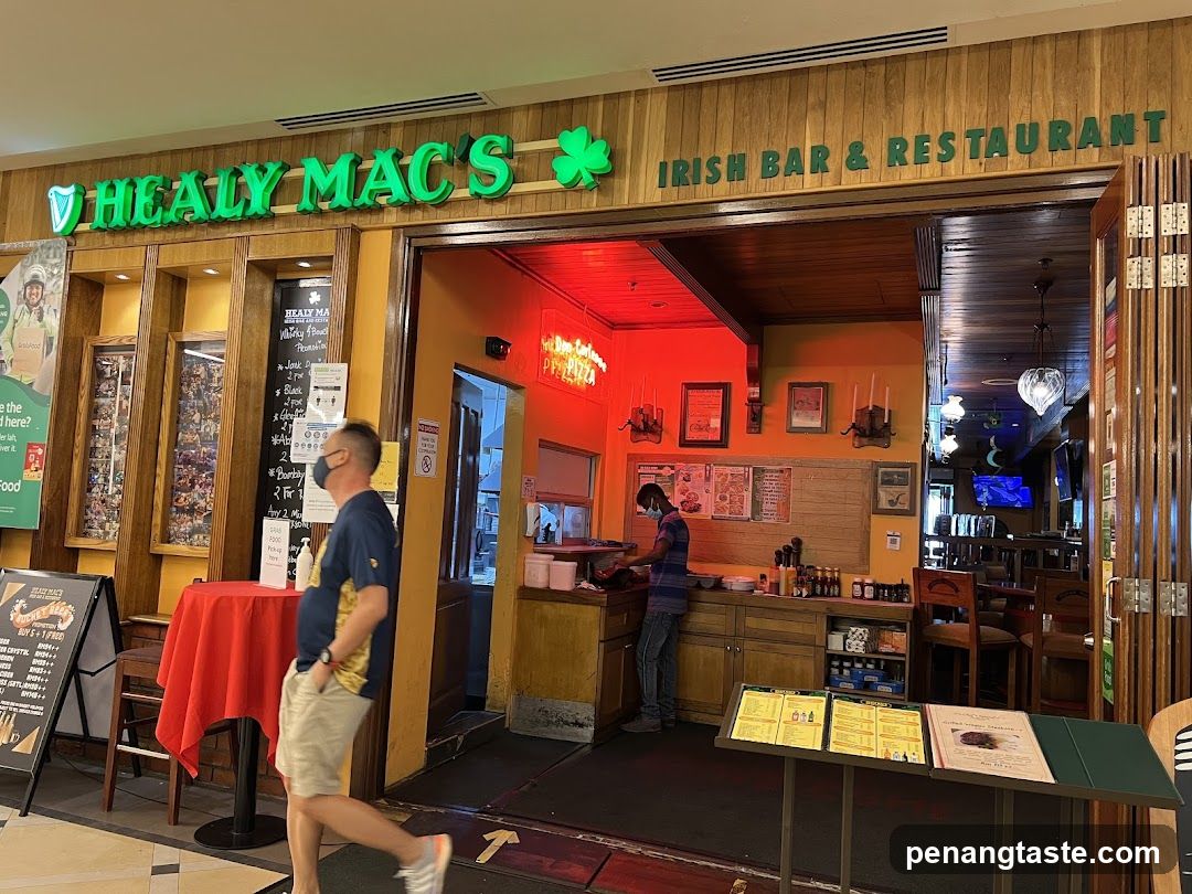 Exterior view of Healy Mac's Irish Bar & Restaurant, featuring green neon signage with shamrock logo above the entrance. Wooden paneling on facade. Red tablecloth-covered table outside. Menu boards inside and near entrance. Staff visible behind counter under red lighting.