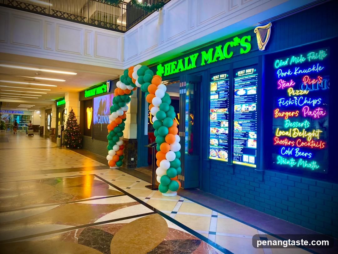 A storefront of Healy Mac's, an Irish pub with green neon signage and a harp logo. The entrance is decorated with orange, white, and green balloons forming an archway. A large illuminated menu board lists items like 'Cord Mole Faile', 'Pork Knuckle', 'Steak Pie', 'Pizza', 'Fish & Chips', 'Burgers', 'Pasta', 'Desserts', 'Local Delight', 'Shooters', 'Cocktails', 'Cold Beers', and 'Stinty Moth'.