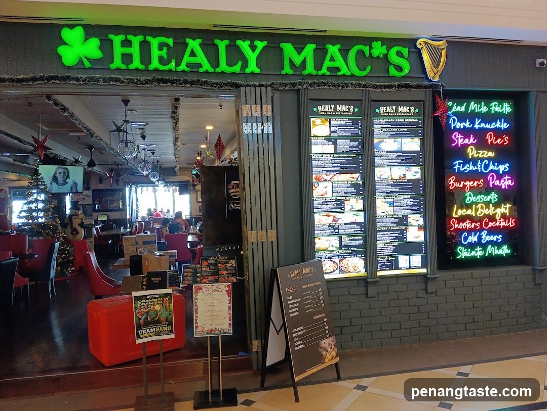 The image shows the exterior of Healy Mac's, a restaurant with green neon signage featuring shamrocks and harp symbols. The entrance is open, revealing an interior with red seating, chandeliers, and Christmas decorations including a tree and hanging ornaments. A large menu board displays various food items like pizza, burgers, pasta, and drinks. There are also promotional posters and flyers near the entrance.