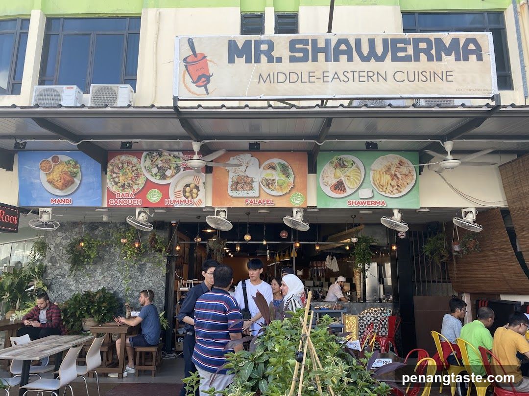A bustling street-level view of Mr. Shawerma, a Middle Eastern eatery with an open-front design. The facade features a large signboard displaying the restaurant name and cuisine type. Below, colorful banners showcase various dishes like Mandi, Daba Gansogh, Kummus, Falafel, and Shawarma. Patrons are seated at outdoor tables, enjoying meals in a casual atmosphere.