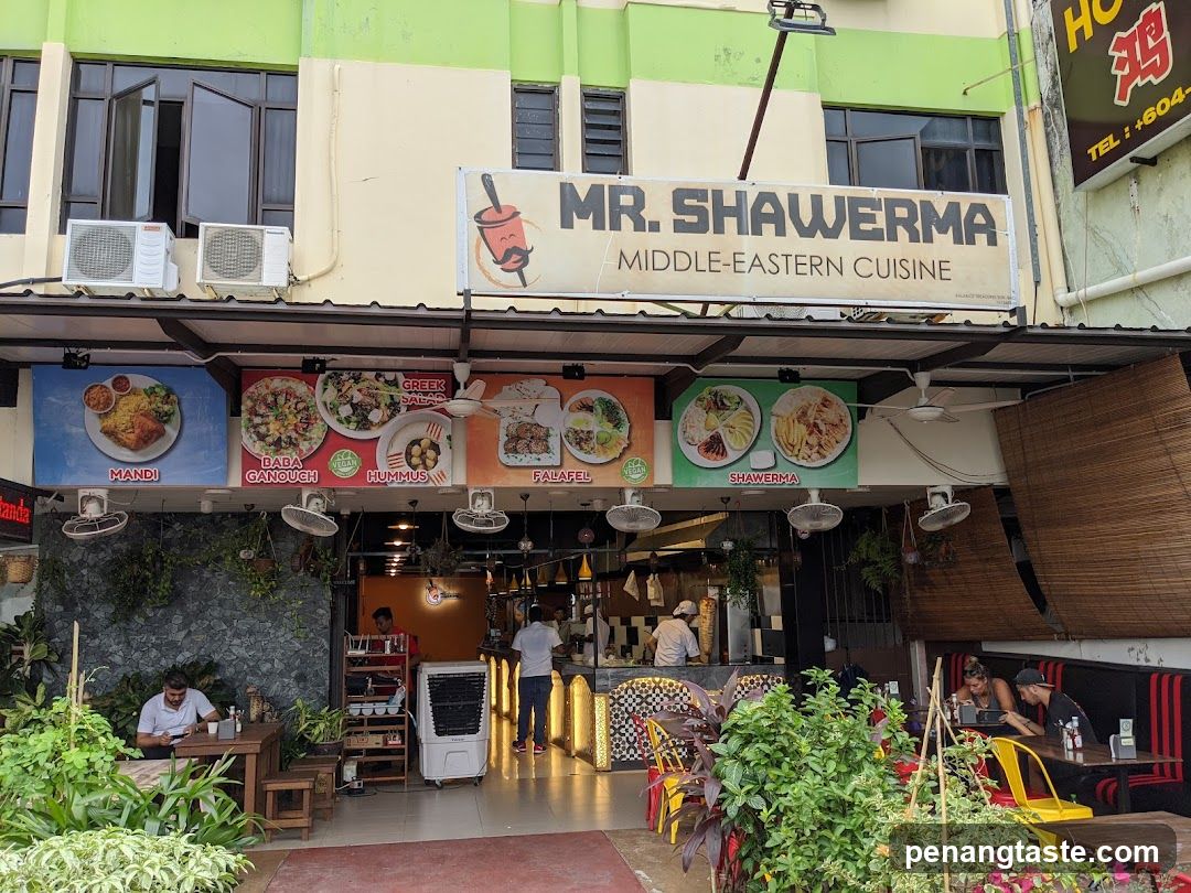 The image shows the exterior of Mr. Shawerma, a restaurant specializing in Middle Eastern cuisine. The shop has a prominent signboard with the name and type of food served. Below the main sign, there are several menu boards displaying various dishes like Mandi, Baba Ghanoush, Koftas, Falafel, and Shawarma. The entrance is open, revealing some interior elements such as staff members working behind the counter. There are also outdoor seating areas visible on either side of the entrance.
