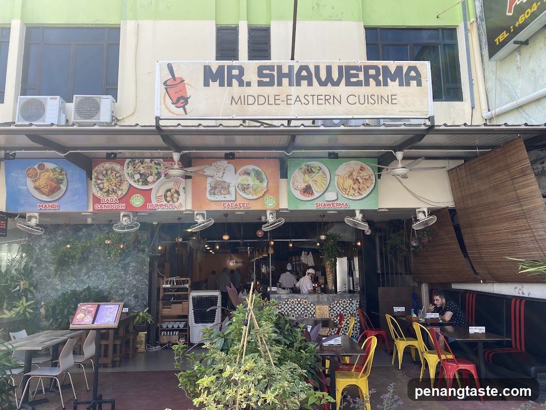 The image shows the exterior of Mr. Shawerma, a restaurant specializing in Middle Eastern cuisine. The storefront features a large sign with the name and logo above the entrance. Below the sign, there are four menu boards displaying images of various dishes: Mandi, Baba Ghanoush, Durgas, Falafel, and Shawarma. The interior is partially visible through the open front, showing staff preparing food behind a counter. Outside, there are several tables and chairs set up for customers, along with some greenery and plants. An air conditioning unit is mounted on the wall to the left.