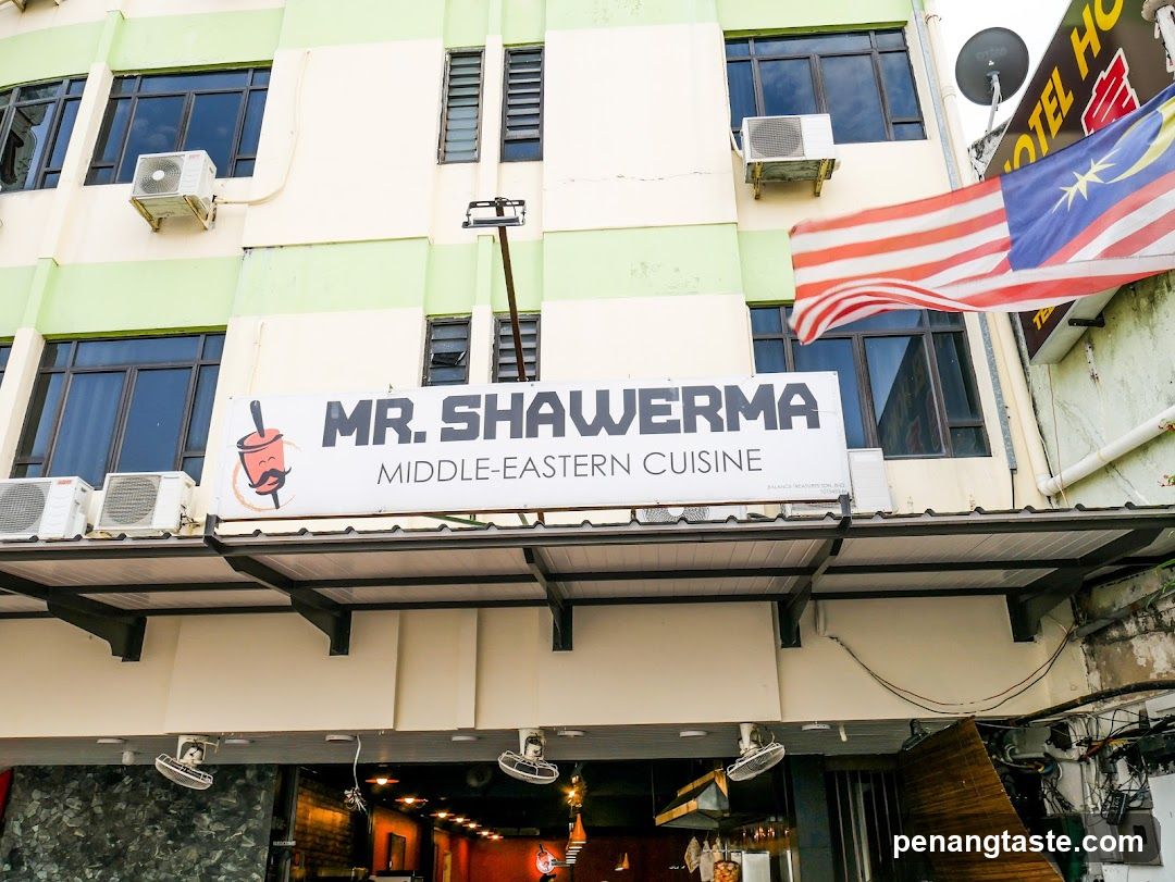 The image shows the exterior of a shop named 'MR. SHAWERMA' specializing in Middle-Eastern Cuisine. The building has multiple air conditioning units mounted on its facade, and there is a Malaysian flag hanging from the side. Below the signboard, the entrance to the restaurant is visible with some interior lighting.