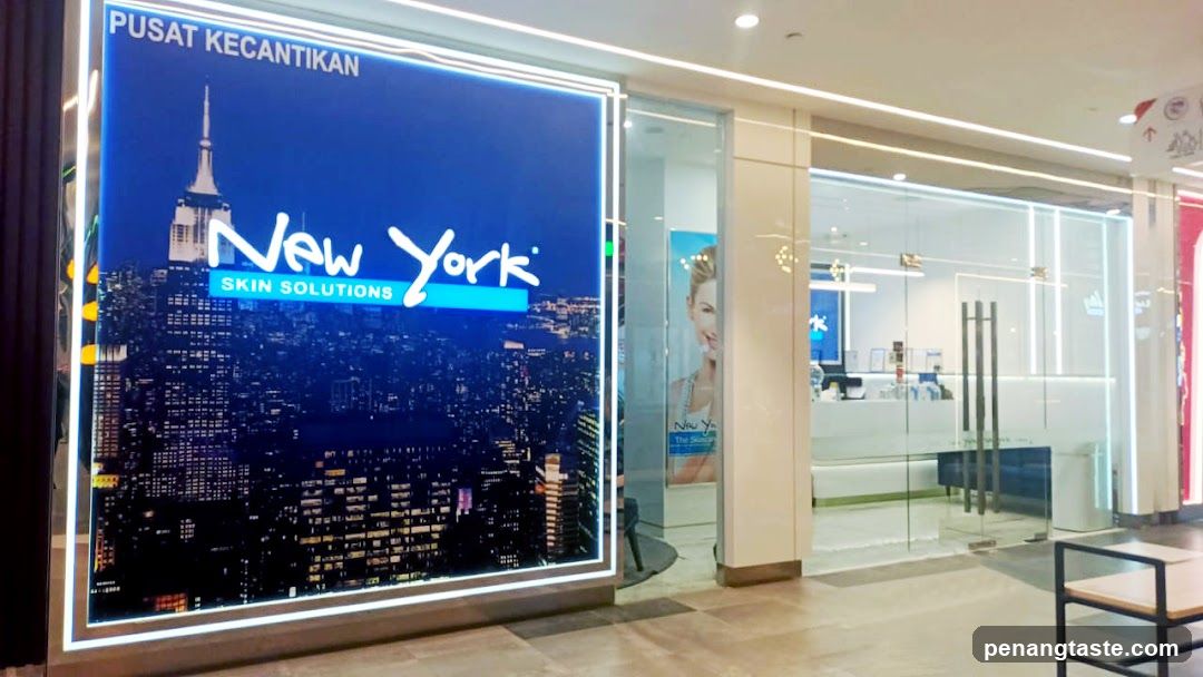 A brightly lit storefront with a large illuminated sign displaying 'PUSAT KECANTIKAN' and 'New York SKIN SOLUTIONS' against a cityscape background. The entrance features glass doors and windows, revealing the interior with promotional posters and modern lighting.