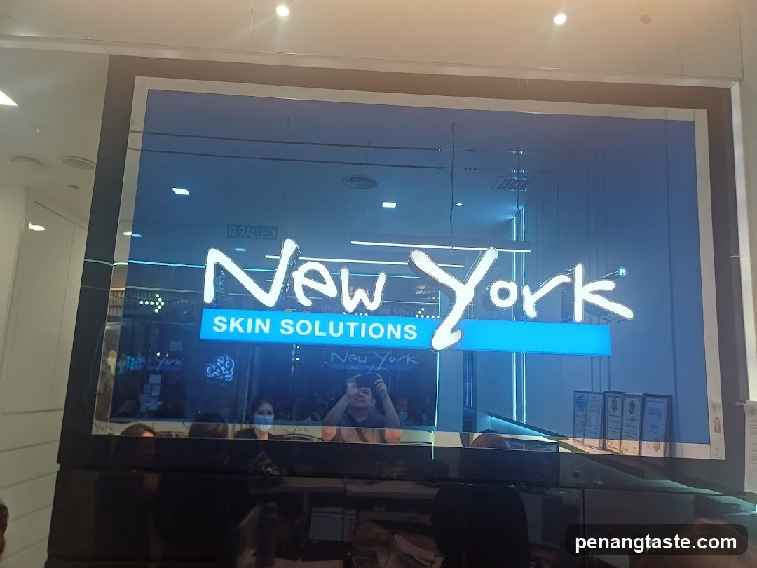 A storefront with a large illuminated sign displaying 'New York' in cursive script above a blue rectangular sign reading 'SKIN SOLUTIONS'. The glass facade reflects interior lighting and people, including someone taking a photo. Framed certificates are visible on the right side of the entrance.