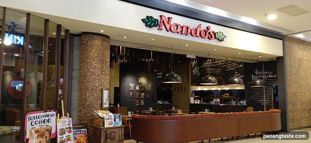 The image shows the storefront of a Nando's restaurant, featuring its prominent red and green logo above the entrance. Inside, there are tables with chairs, hanging pendant lights, and chalkboard menus on the walls displaying various food items. A promotional standee for 'Bottomless Coffee' is visible near the entrance.