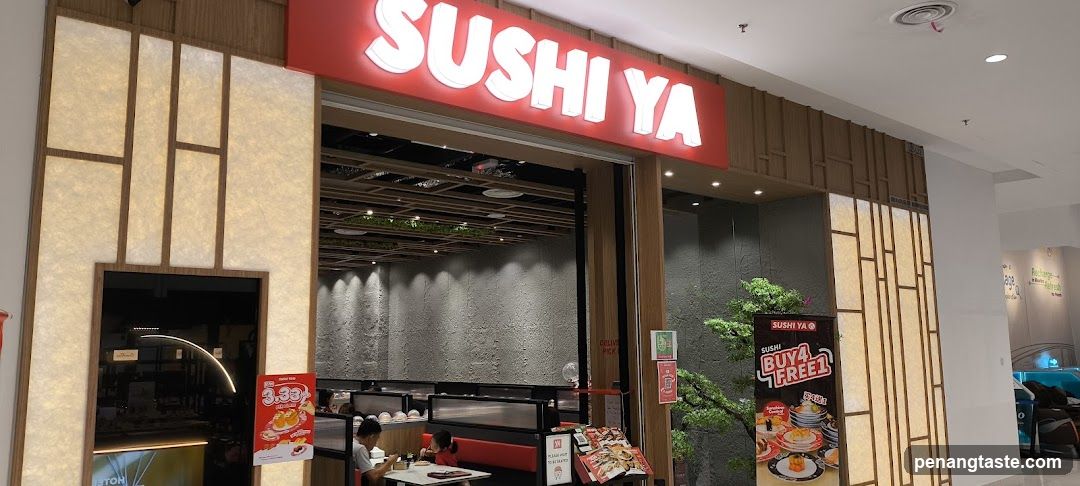 The image shows the exterior entrance of a Sushi Ya restaurant, featuring a prominent red sign with white lettering above the doorway. The facade includes wooden paneling and illuminated accents. Inside, customers are seated at tables in booths. A promotional poster on the right advertises 'Buya Free1', while smaller signs display prices or offers near the entrance.