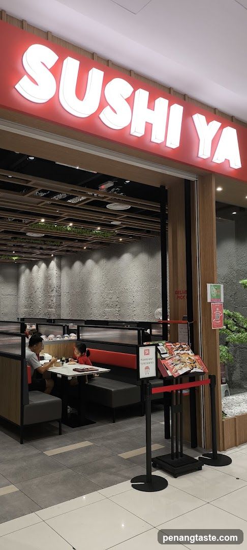 The image shows the entrance of a Sushi Ya restaurant with its illuminated signage prominently displayed above the doorway. Inside, customers are seated at tables in a modern dining area featuring wooden accents and greenery on the ceiling.