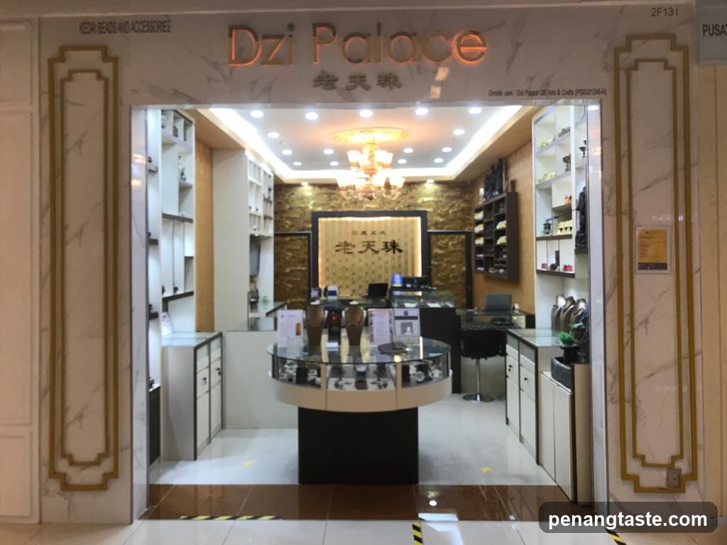 The image shows the storefront of a jewelry shop named 'Dzi Palace' with Chinese characters below it. The entrance is framed by marble columns with gold accents, and the interior features a well-lit display area with shelves on either side holding various items. A central circular counter showcases jewelry pieces under glass. The ceiling has recessed lighting and a chandelier.