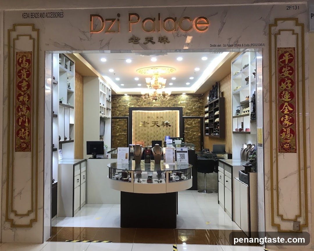 The image shows the storefront of a jewelry shop named 'Dzi Palace' with Chinese characters below it. The entrance is framed by marble columns adorned with vertical red banners featuring gold calligraphy. Inside, there are display cases showcasing necklaces and other jewelry items on mannequin busts. A reception desk is visible in the background along with shelving units holding additional products. The ceiling has recessed lighting and a chandelier.