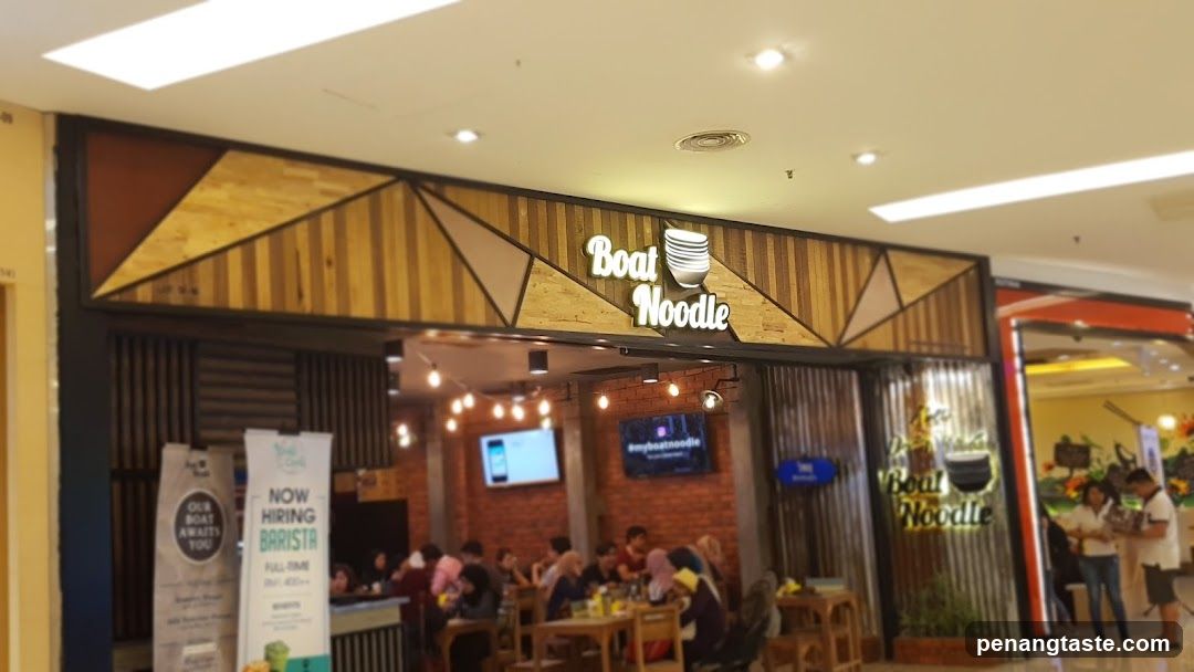 A photo of the physical exterior entrance/storefront facade with visible signage reading 'Boat Noodle' above a wooden-patterned awning. The shop interior is partially visible, showing customers dining at tables and hanging lights.