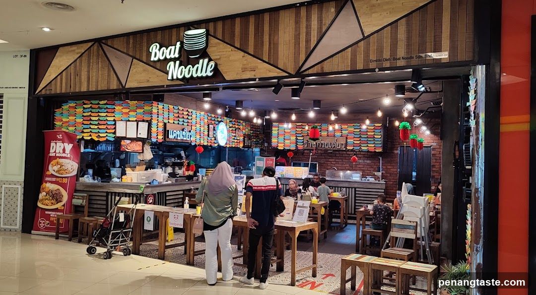 A storefront with wooden paneling and geometric patterns, featuring a sign reading 'Boat Noodle' in illuminated letters. Inside, customers are seated at tables, and colorful decorations hang from the ceiling. A red poster on the left advertises 'DRY RICE'.