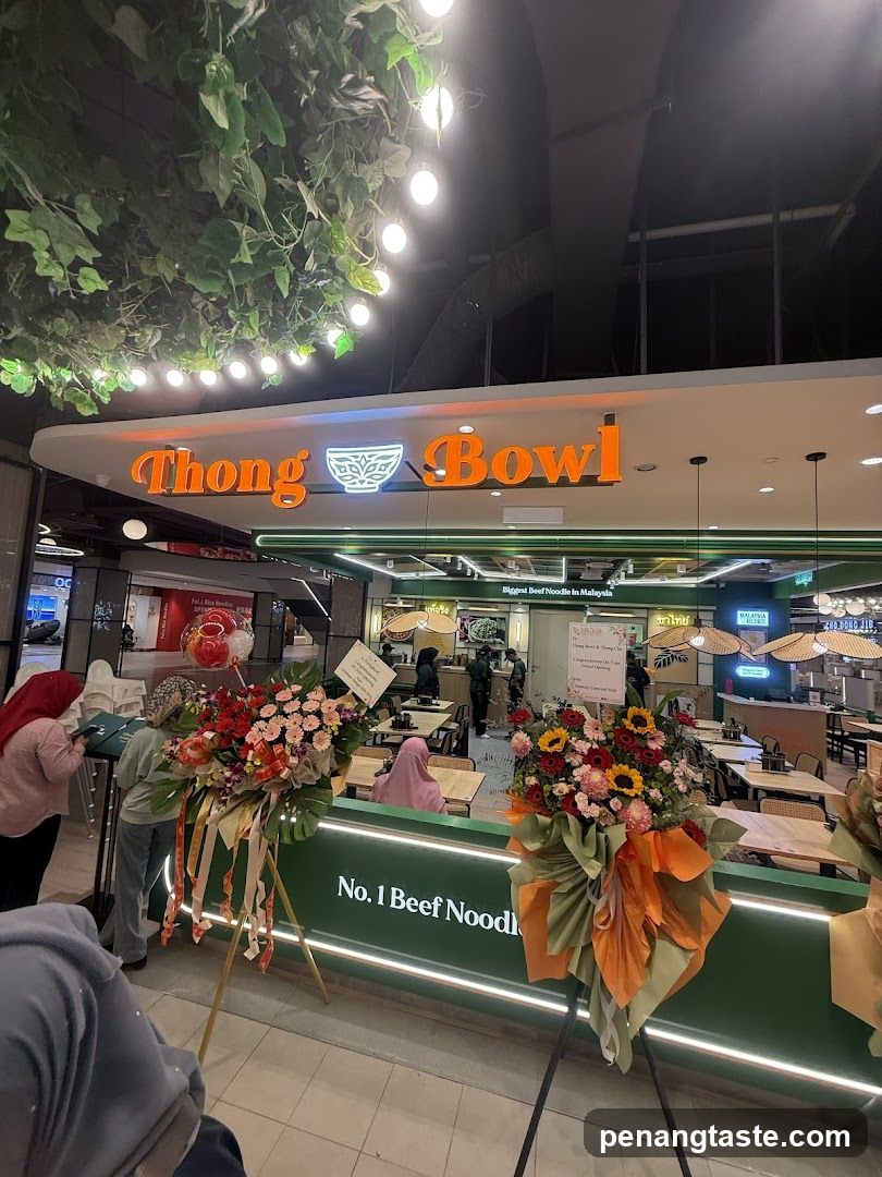 The image shows the storefront of Thong Bowl, a restaurant chain specializing in beef noodles. The sign above the entrance reads 'Thong Bowl' with an illuminated logo featuring a bowl icon. Below the sign, there are floral arrangements on stands and people standing near the entrance. Inside, customers can be seen seated at tables.