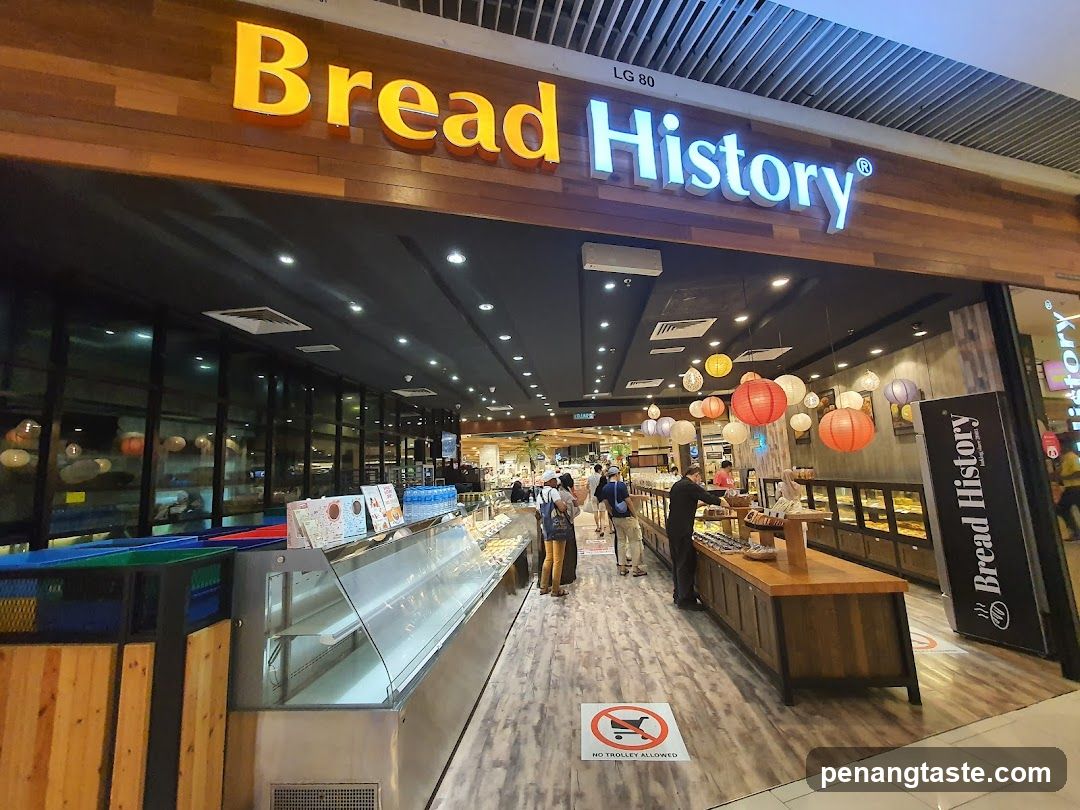 The image shows the exterior entrance of a Bread History store, featuring prominent signage with illuminated 'Bread' in yellow and 'History' in white letters mounted on a wooden facade. Inside, customers browse various bread products displayed under glass cases and hanging lanterns decorate the ceiling area.