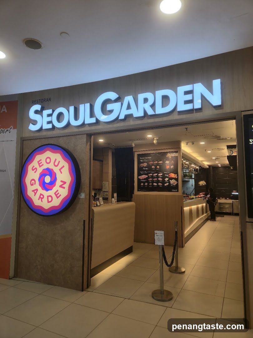 The image shows the storefront of a restaurant named 'SEOULGARDEN'. The name is prominently displayed in large, illuminated white letters on the facade above the entrance. To the left of the entrance, there is a circular sign with the same name and a colorful spiral design inside. Inside, through the open doorway, a counter area is visible along with a menu board displaying various food items. There are also stanchions with ropes guiding customers near the entrance.